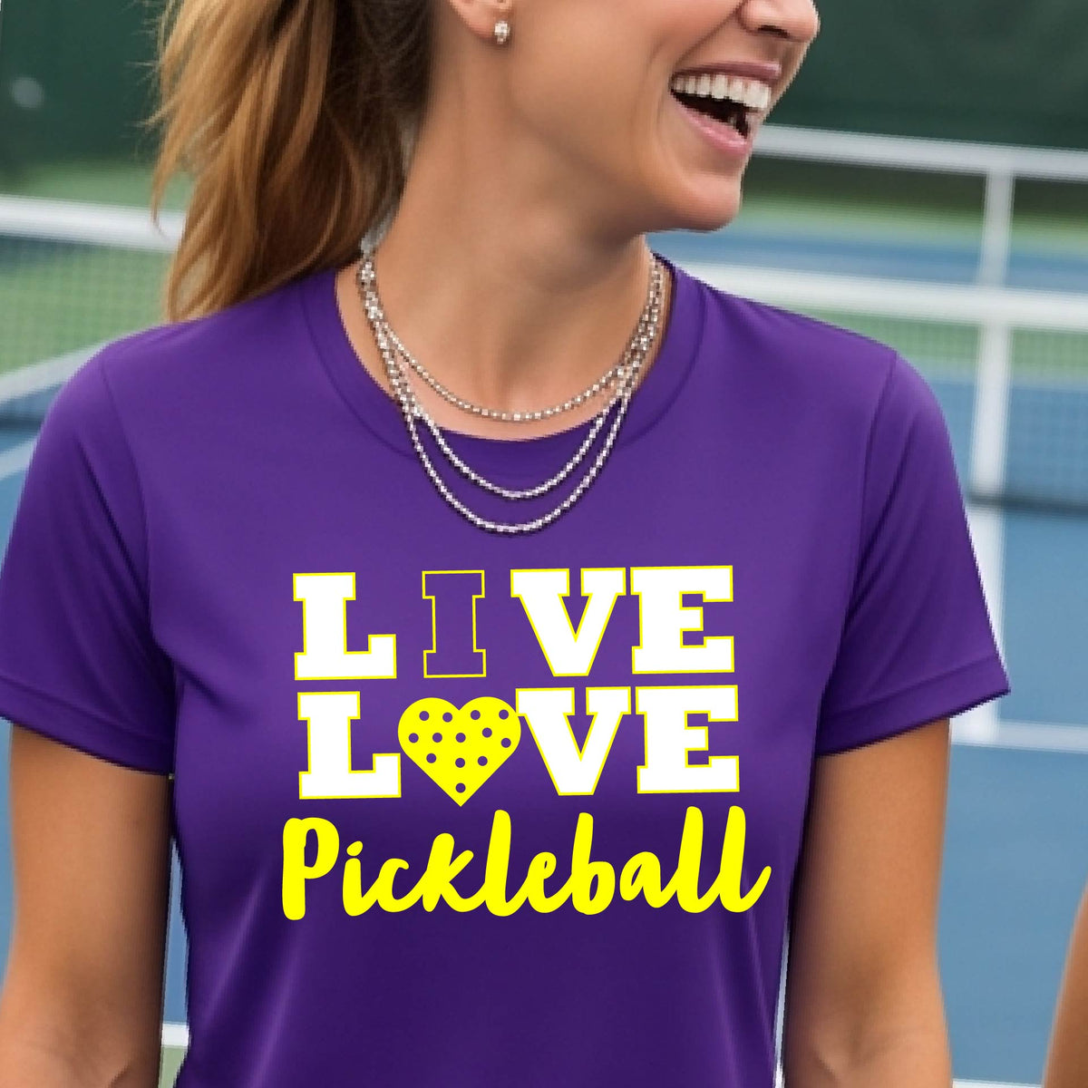 A woman on a pickleball court wearing a purple short-sleeve t-shirt with a white and yellow graphic that reads 'LIVE LOVE Pickleball' and a heart-shaped pickleball dot pattern inside the word 'LOVE'.