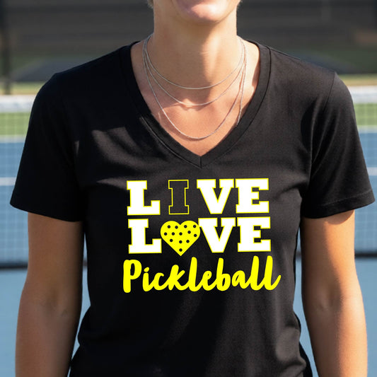 A smiling woman on a pickleball court wearing a black V-neck t-shirt with a yellow graphic that reads 'LIVE LOVE Pickleball' with a heart-shaped pickleball motif.