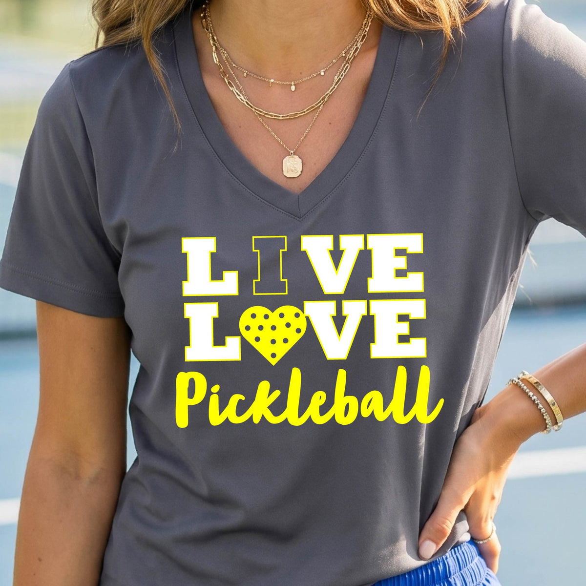 A smiling woman on a pickleball court wearing a dark gray V-neck t-shirt with a yellow graphic that reads 'LIVE LOVE Pickleball' with a heart-shaped pickleball motif.