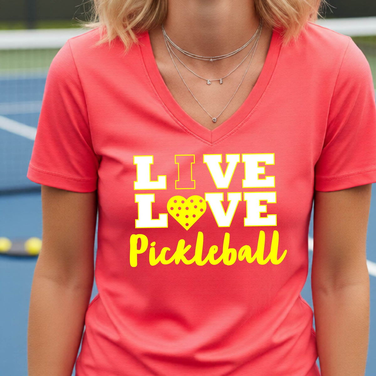 A smiling woman on a pickleball court wearing a CORAL PINK V-neck t-shirt with a yellow graphic that reads 'LIVE LOVE Pickleball' with a heart-shaped pickleball motif.
