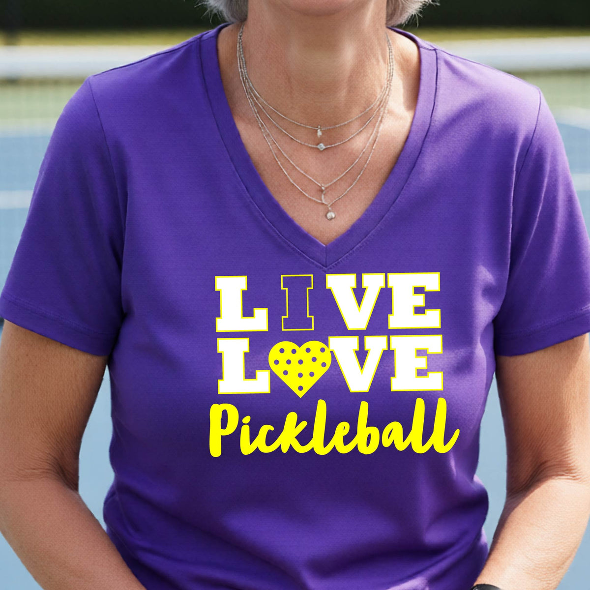 A smiling woman on a pickleball court wearing a purple V-neck t-shirt with a yellow graphic that reads 'LIVE LOVE Pickleball' with a heart-shaped pickleball motif.