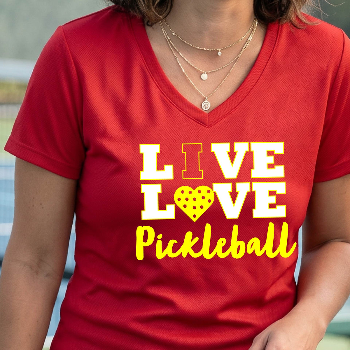 A smiling woman on a pickleball court wearing a red V-neck t-shirt with a yellow graphic that reads 'LIVE LOVE Pickleball' with a heart-shaped pickleball motif.