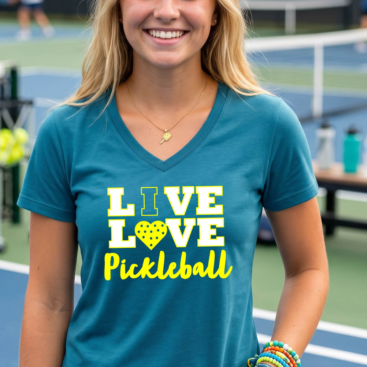 A smiling woman on a pickleball court wearing a teal blue V-neck t-shirt with a yellow graphic that reads 'LIVE LOVE Pickleball' with a heart-shaped pickleball motif.