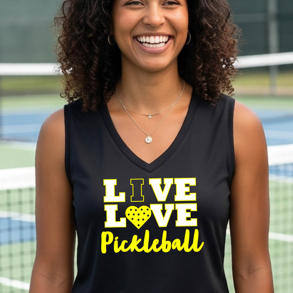 A woman on a pickleball court wearing a black sleeveless tank top with the text 'LIVE LOVE Pickleball' printed in white and yellow. The background shows a blurred pickleball court net and outdoor setting.