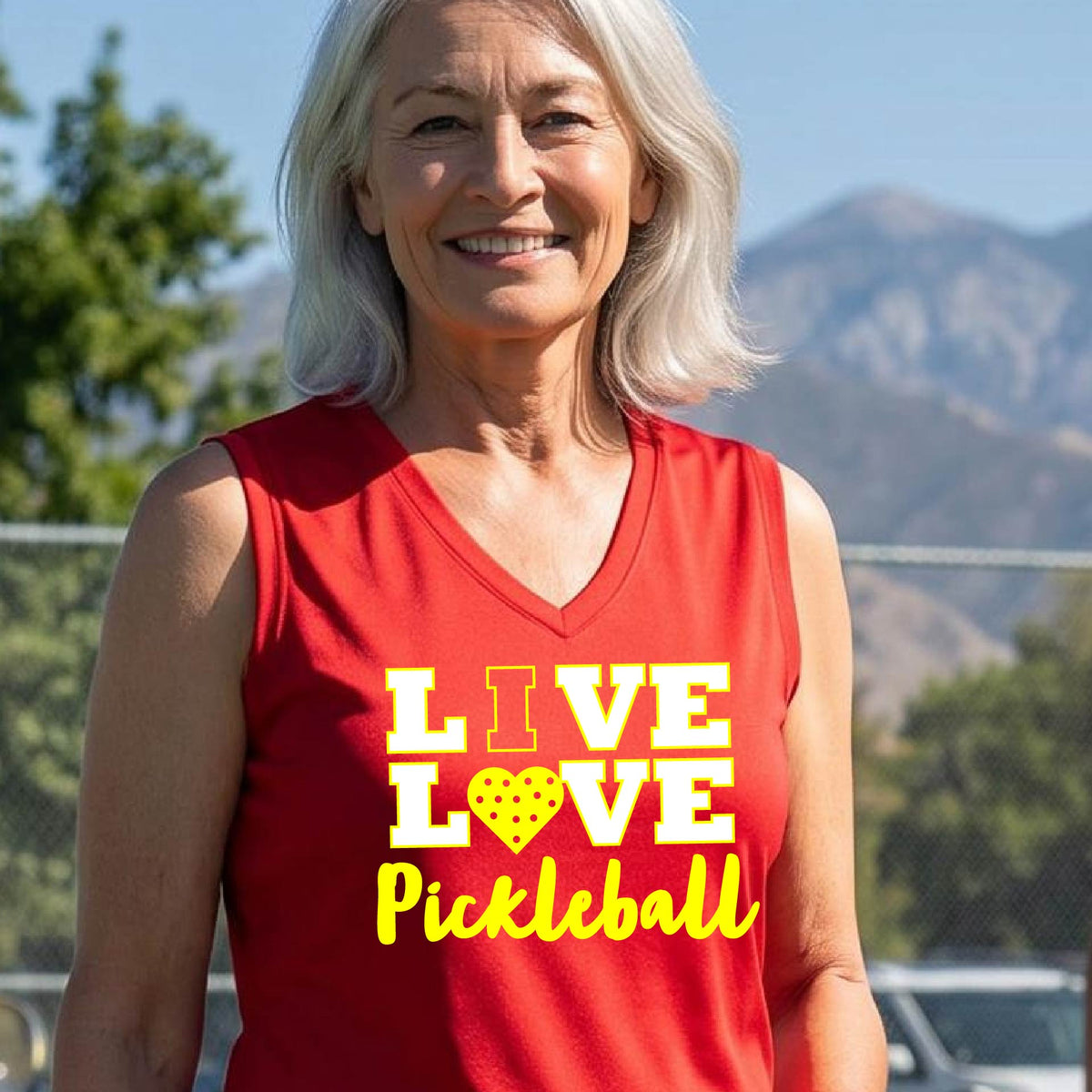 A woman on a pickleball court wearing a red sleeveless tank top with the text 'LIVE LOVE Pickleball' printed in white and yellow. The background shows a blurred pickleball court net and outdoor setting.