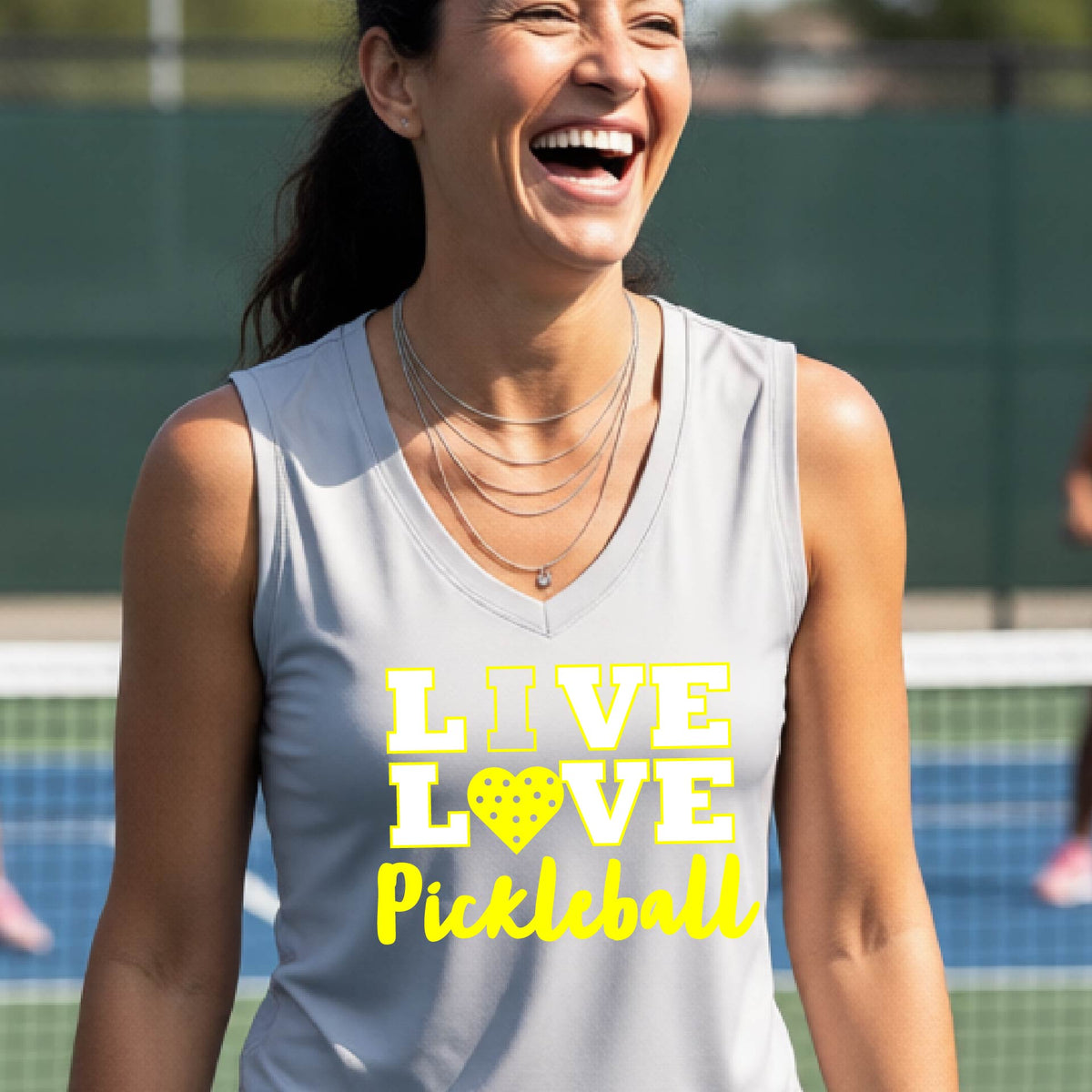 A woman on a pickleball court wearing a silver gray sleeveless tank top with the text 'LIVE LOVE Pickleball' printed in white and yellow. The background shows a blurred pickleball court net and outdoor setting.