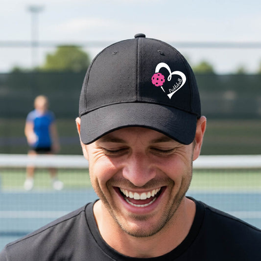 A close up of an outdoor pickleball court and a black baseball cap with a white heart outline, a pink pickleball graphic, and the text 'Pickleball'.