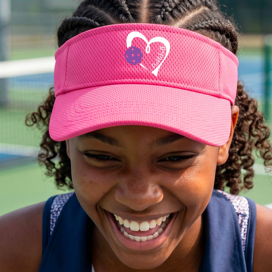 A close-up lifestyle shot of a smiling woman on a pickleball court wearing a bright pink visor with a white heart and purple pickleball, with the text 'pickleball' inside the heart, graphic on the front.