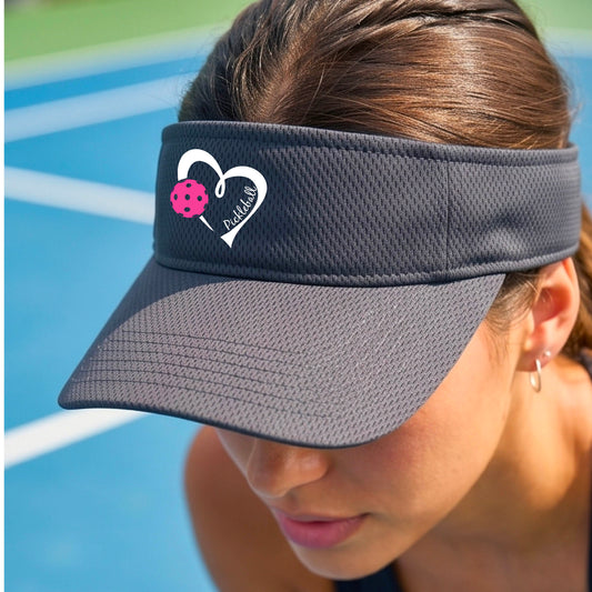 A close-up of a dark gray athletic visor with the graphic features of a white heart with a pickleball icon and the wrod 'pickleball' written inside.