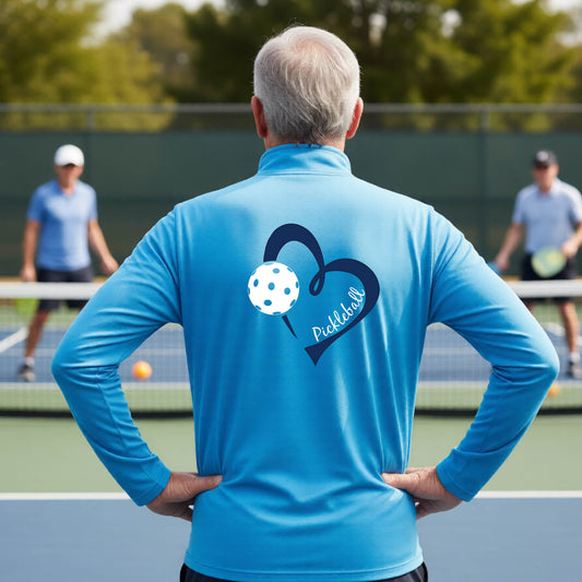 A man viewed from behind on an outdoor pickleball court, wearing a light blue long-sleeve performance quarter-zip shirt with a navy blue heart-shaped graphic and a white pickleball on the back, alongside the script text 'Pickleball'.