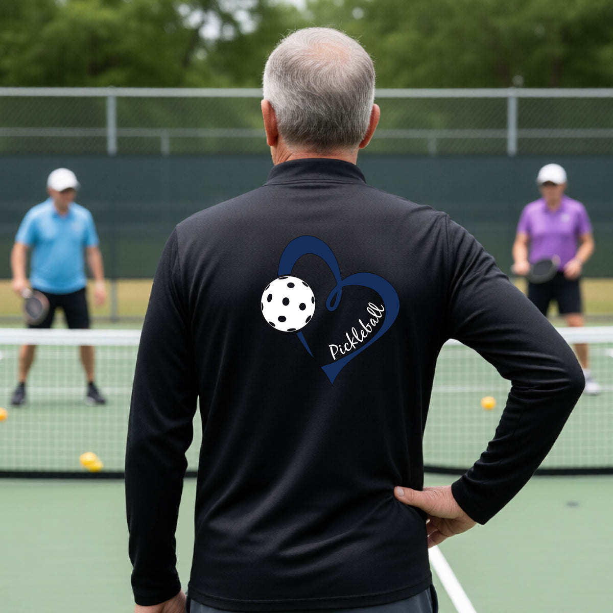 A man viewed from behind on an outdoor pickleball court, wearing a black long-sleeve performance quarter-zip shirt with a navy blue heart-shaped graphic and a white pickleball on the back, alongside the script text 'Pickleball'.