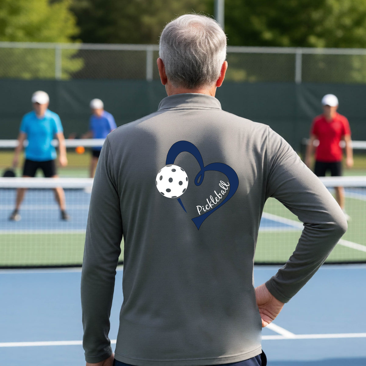 A man viewed from behind on an outdoor pickleball court, wearing a gray long-sleeve performance quarter-zip shirt with a navy blue heart-shaped graphic and a white pickleball on the back, alongside the script text 'Pickleball'.