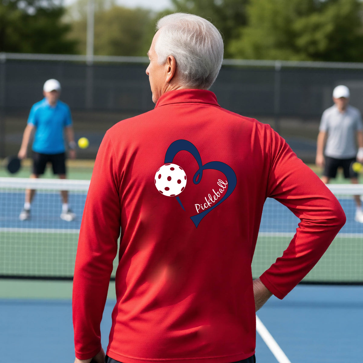 A man viewed from behind on an outdoor pickleball court, wearing a red long-sleeve performance quarter-zip shirt with a navy blue heart-shaped graphic and a white pickleball on the back, alongside the script text 'Pickleball'.