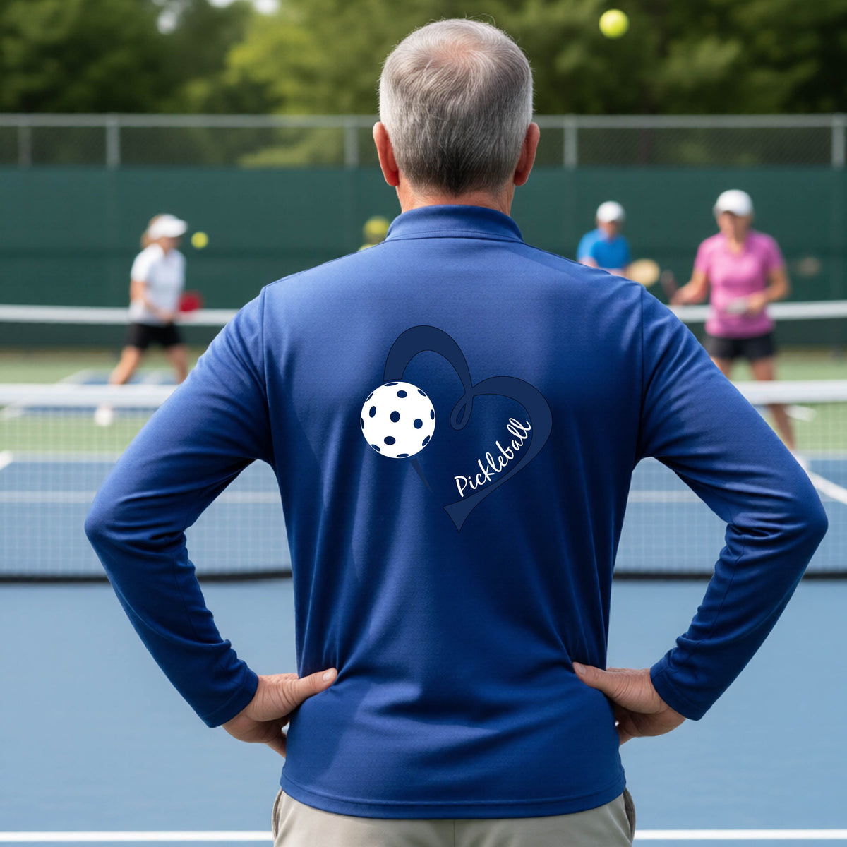 A man viewed from behind on an outdoor pickleball court, wearing a royal blue long-sleeve performance quarter-zip shirt with a navy blue heart-shaped graphic and a white pickleball on the back, alongside the script text 'Pickleball'.