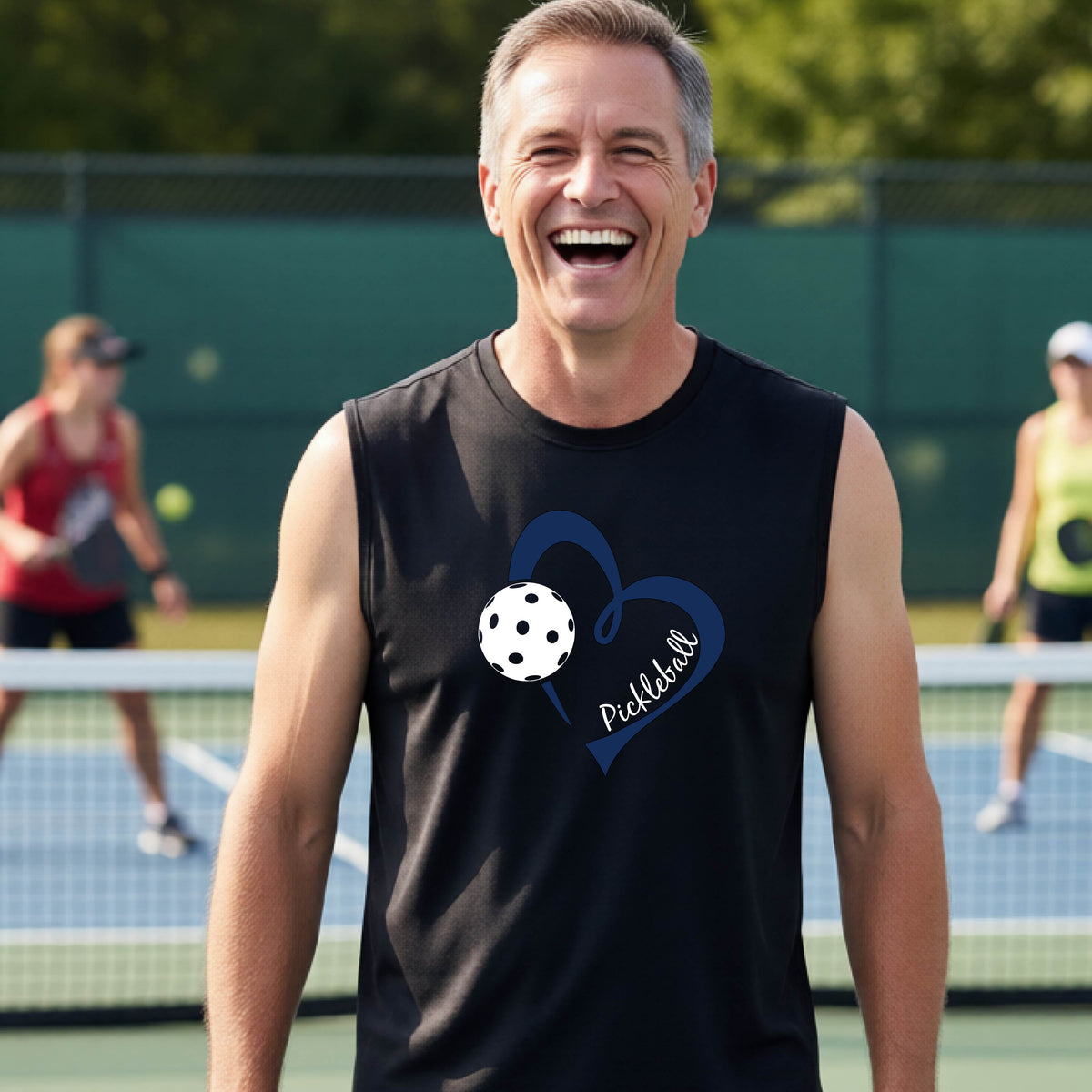 A smiling man on a pickleball court wearing a black sleeveless shirt with a navy heart, pickleball graphic, and the word 'Pickleball' printed on the front.