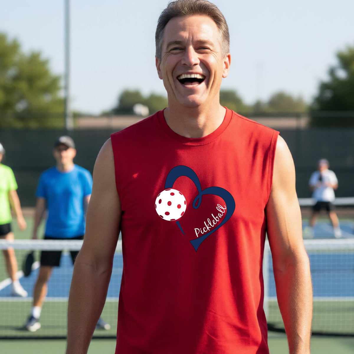 A smiling man on a pickleball court wearing a red sleeveless shirt with a navy heart, pickleball graphic, and the word 'Pickleball' printed on the front.