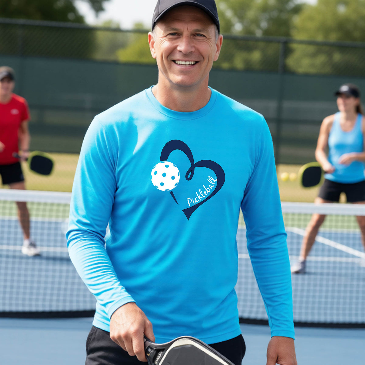 A smiling man on a pickleball court wearing a bright blue long-sleeve athletic shirt with a blue heart, pickleball, and the word 'Pickleball' printed on the front.