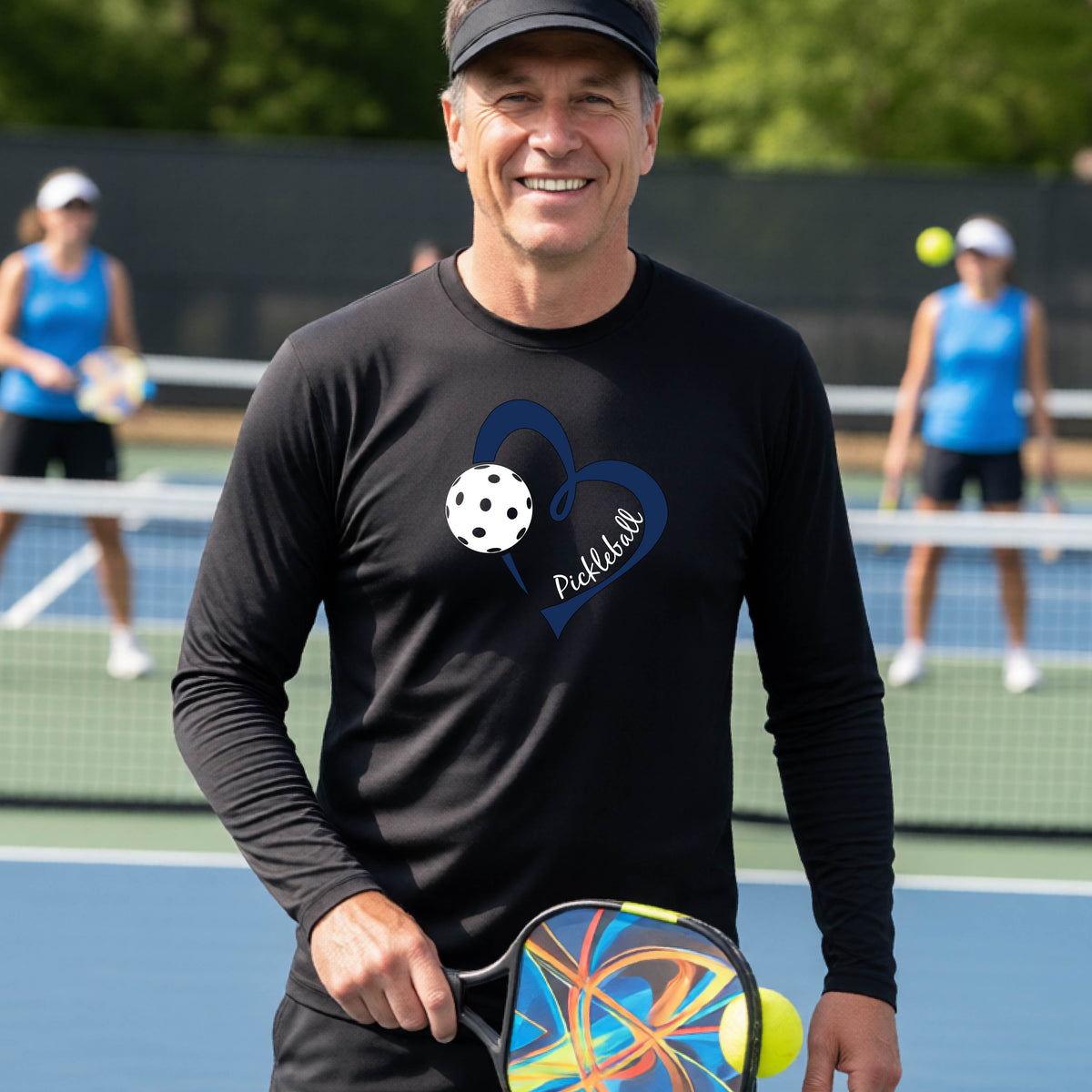 A smiling man on a pickleball court wearing a black long-sleeve athletic shirt with a blue heart, pickleball, and the word 'Pickleball' printed on the front.