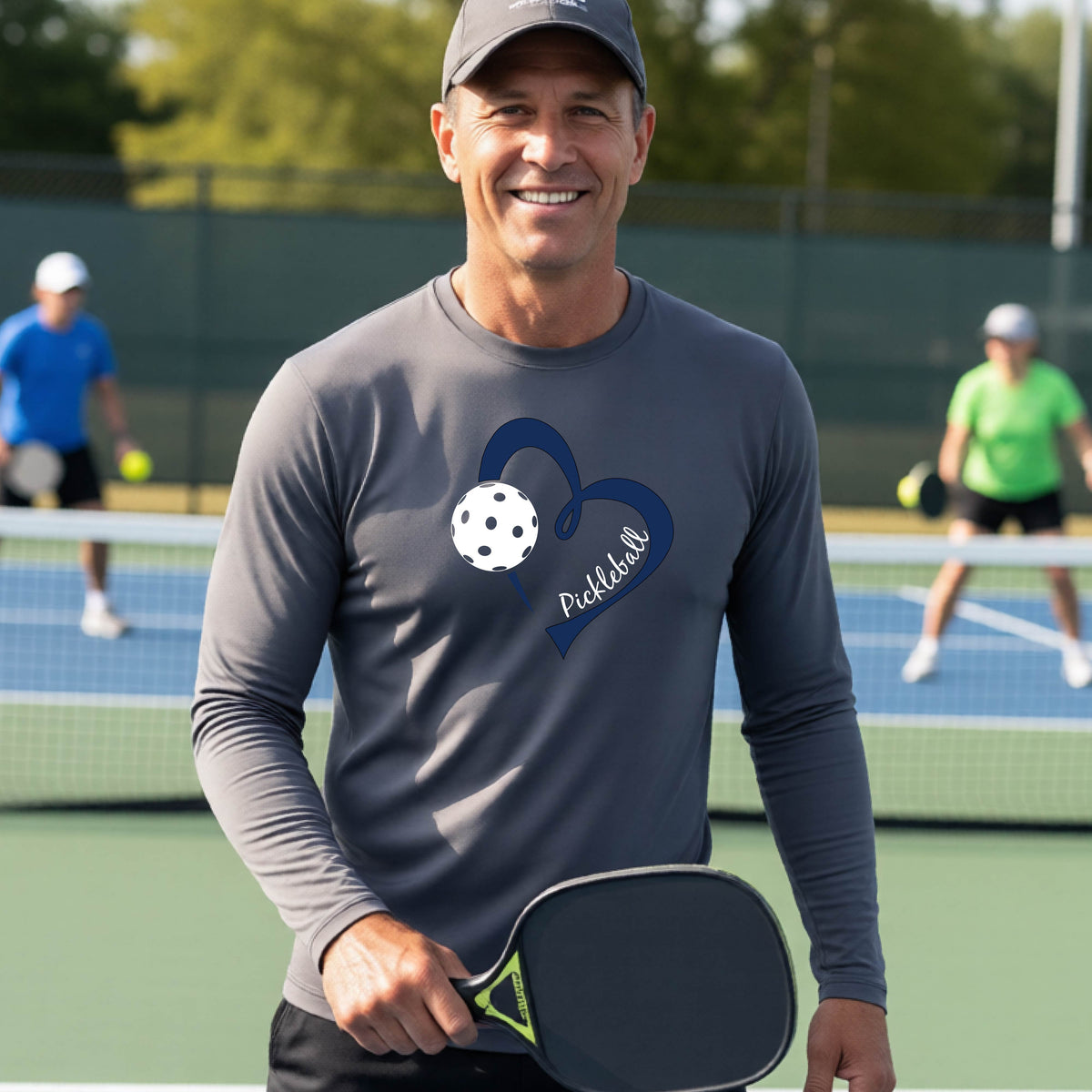 A smiling man on a pickleball court wearing a dark gray long-sleeve athletic shirt with a blue heart, pickleball, and the word 'Pickleball' printed on the front.