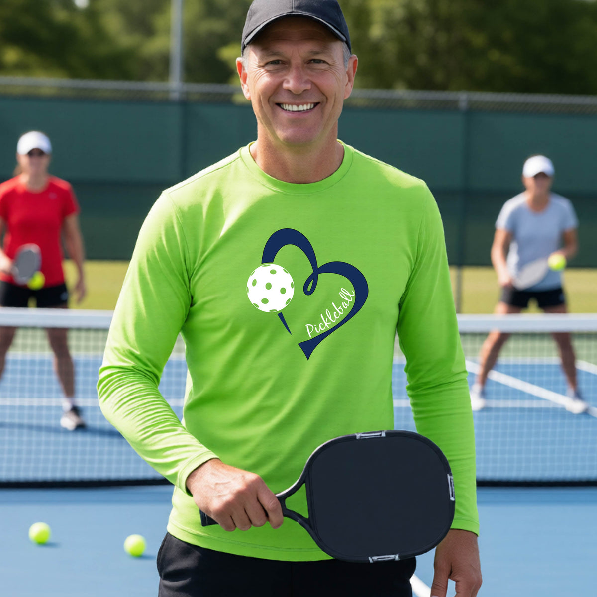 A smiling man on a pickleball court wearing a lime green long-sleeve athletic shirt with a blue heart, pickleball, and the word 'Pickleball' printed on the front.