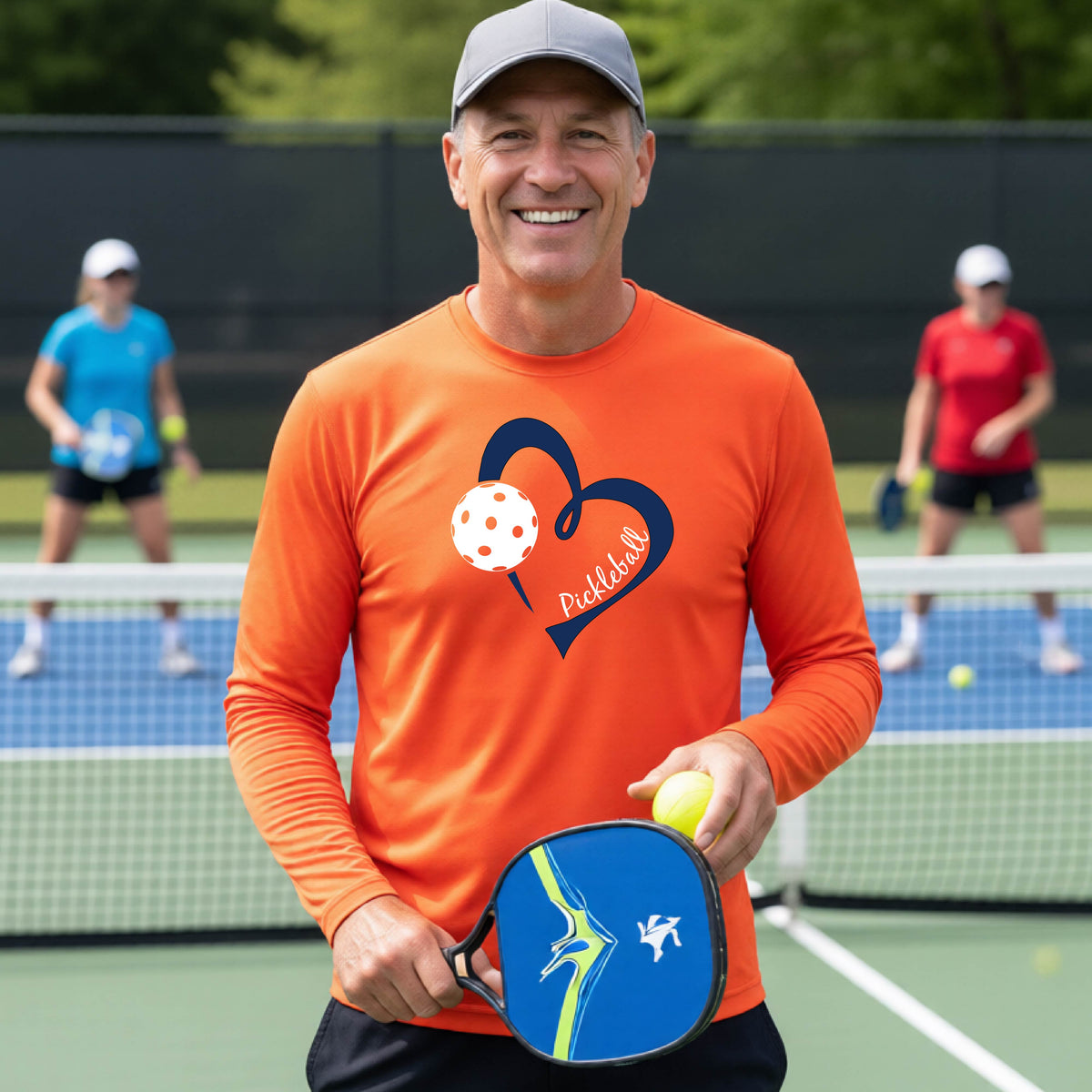 A smiling man on a pickleball court wearing a bright orange long-sleeve athletic shirt with a blue heart, pickleball, and the word 'Pickleball' printed on the front.