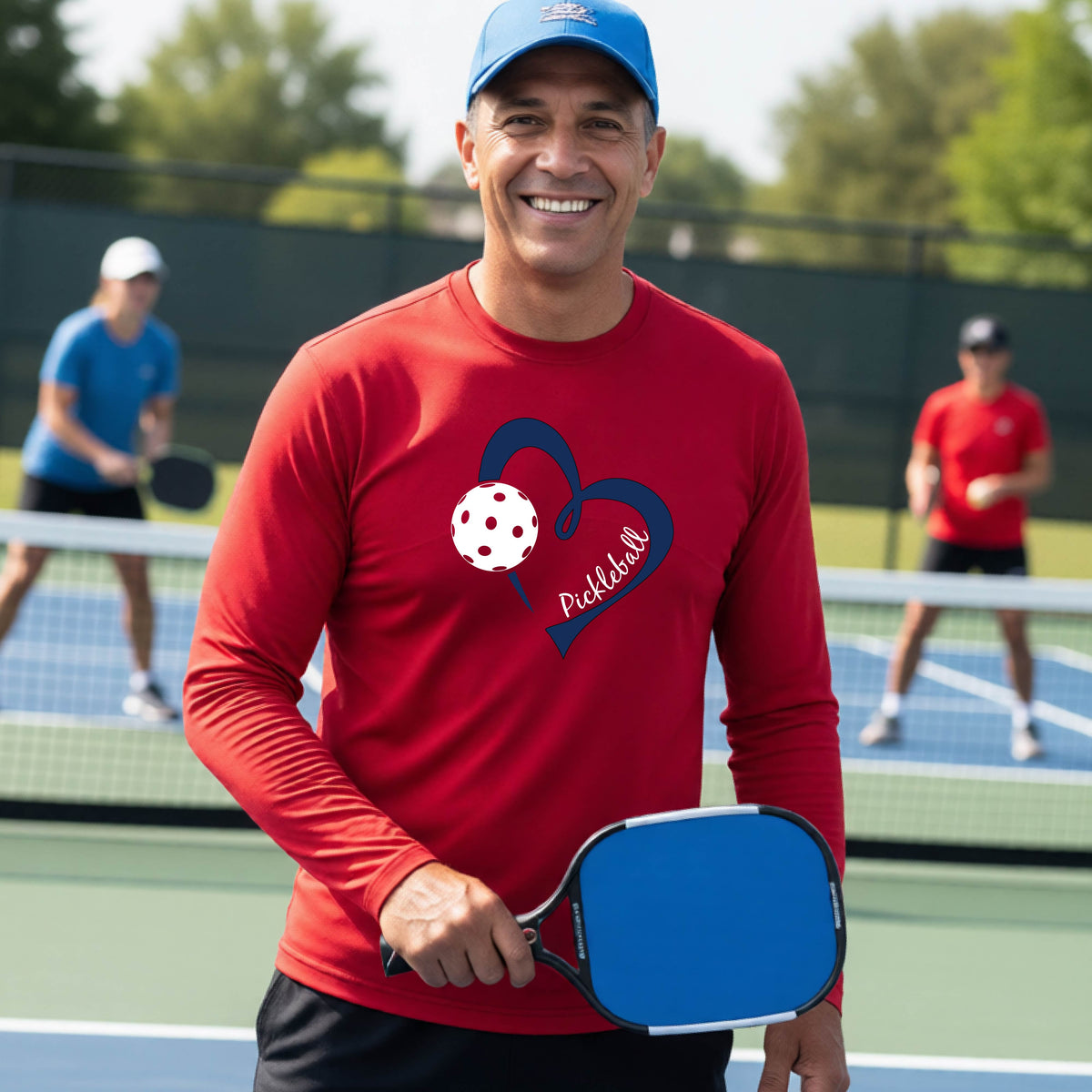 A smiling man on a pickleball court wearing a red long-sleeve athletic shirt with a blue heart, pickleball, and the word 'Pickleball' printed on the front.