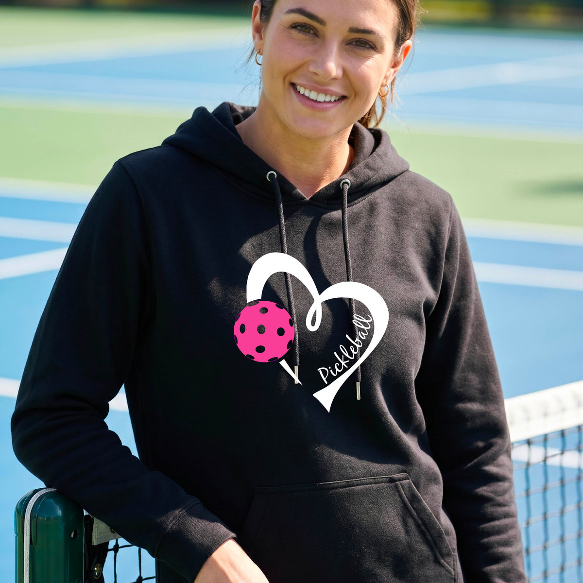 A smiling man wearing a black hoodie with a white heart outline, a pink pickleball graphic, and the word 'Pickleball' printed on the front, standing on an outdoor pickleball court.