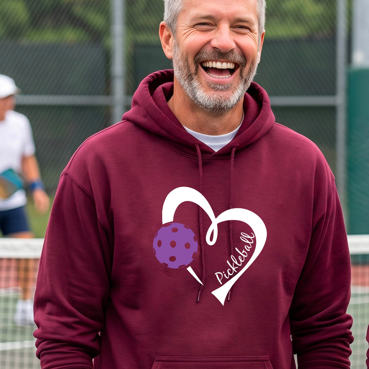 A smiling man wearing a maroon hoodie with a white heart outline graphic and a purple picklkeball design and the texgt 'Pickleball' on the chest, standing on a pickleball court.