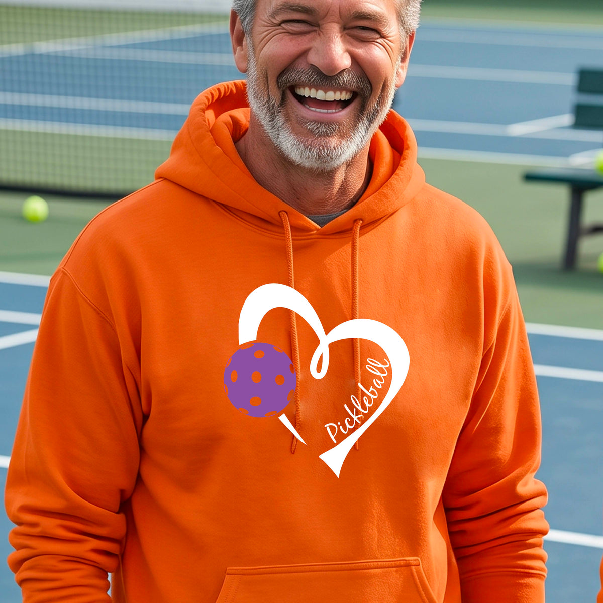 A smiling man wearing an orange hoodie with a white heart outline graphic and a purple picklkeball design and the texgt 'Pickleball' on the chest, standing on a pickleball court.