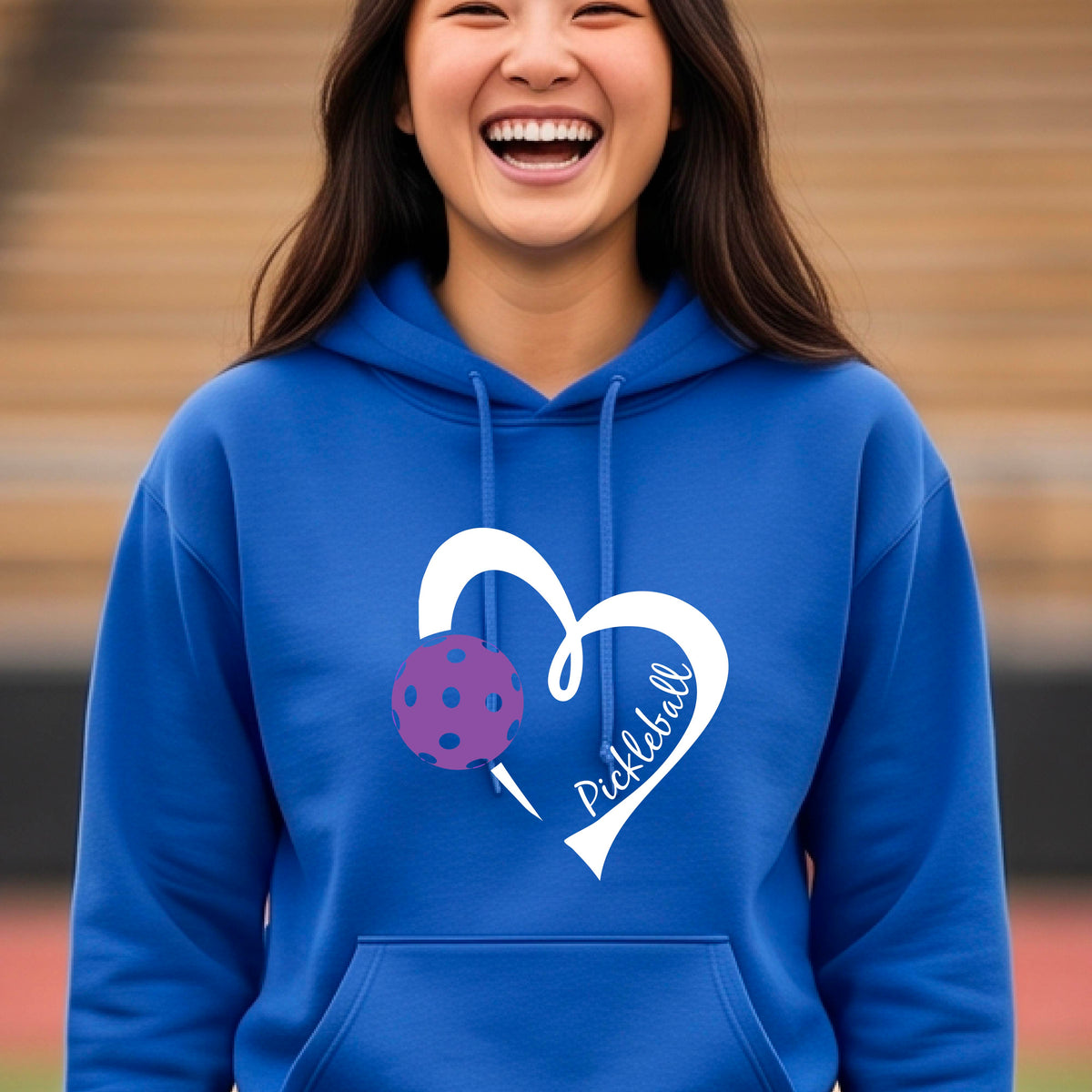 A smiling man wearing a royal blue hoodie with a white heart outline graphic and a purple picklkeball design and the texgt 'Pickleball' on the chest, standing on a pickleball court.