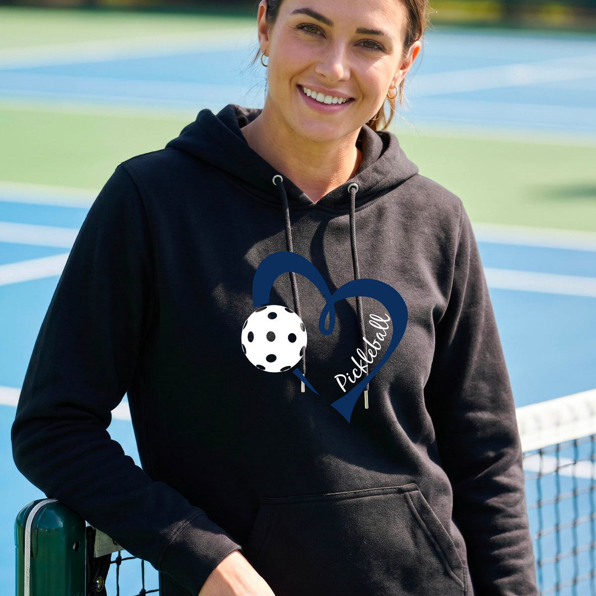 A smiling woman wearing a black pullover hoodie with a navy blue heart outline, a white pickleball graphic, and the word 'Pickleball' on the front.