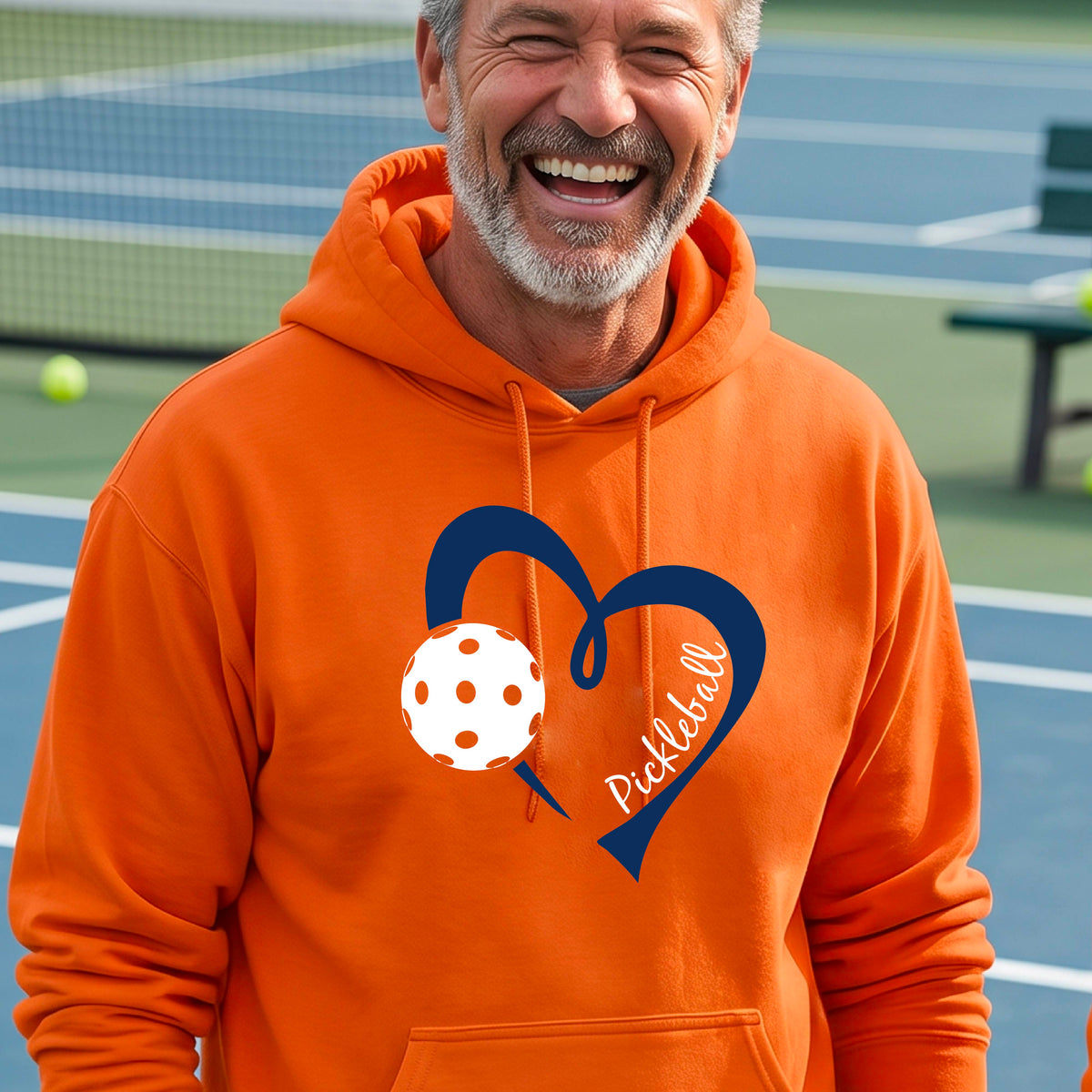 A smiling man wearing an orange pullover hoodie with a navy blue heart outline, a white pickleball graphic, and the word 'Pickleball' on the front.