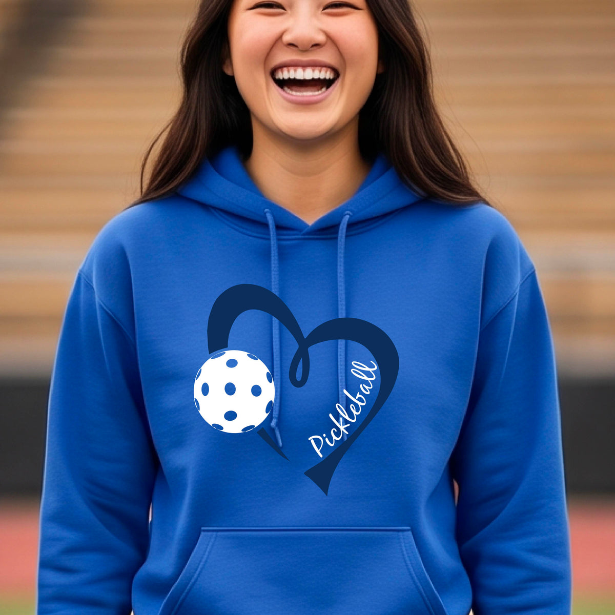 A smiling woman wearing a royal blue pullover hoodie with a navy blue heart outline, a white pickleball graphic, and the word 'Pickleball' on the front.