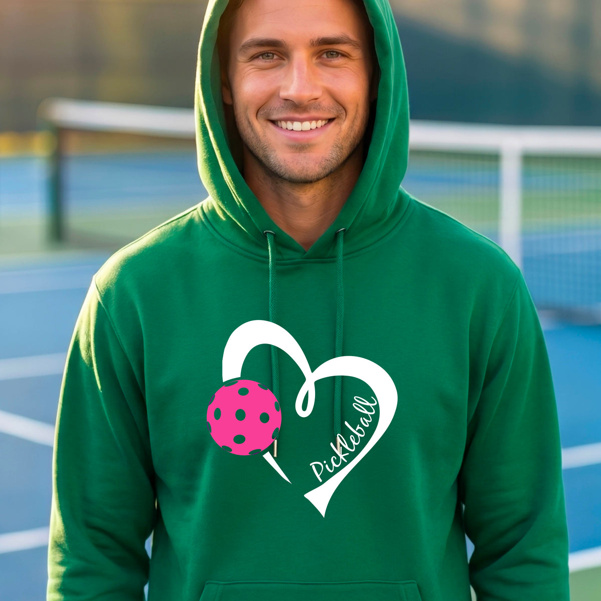 A smiling man wearing a green hoodie with a white heart outline, a pink pickleball graphic, and the word 'Pickleball' printed on the front, standing on an outdoor pickleball court.