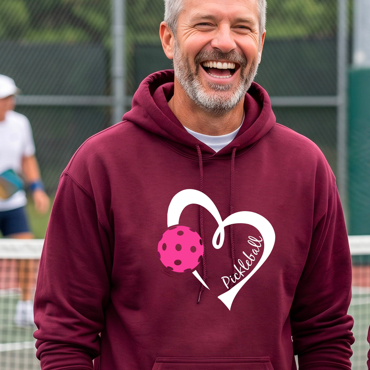A smiling man wearing a maroon hoodie with a white heart outline, a pink pickleball graphic, and the word 'Pickleball' printed on the front, standing on an outdoor pickleball court.