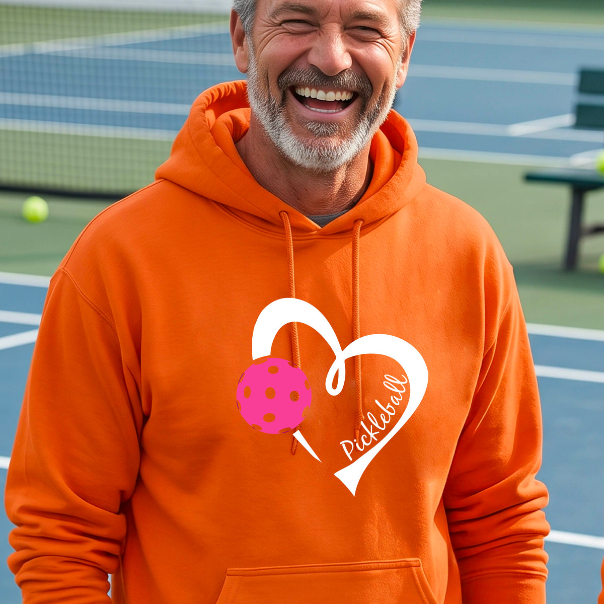 A smiling man wearing an orange hoodie with a white heart outline, a pink pickleball graphic, and the word 'Pickleball' printed on the front, standing on an outdoor pickleball court.