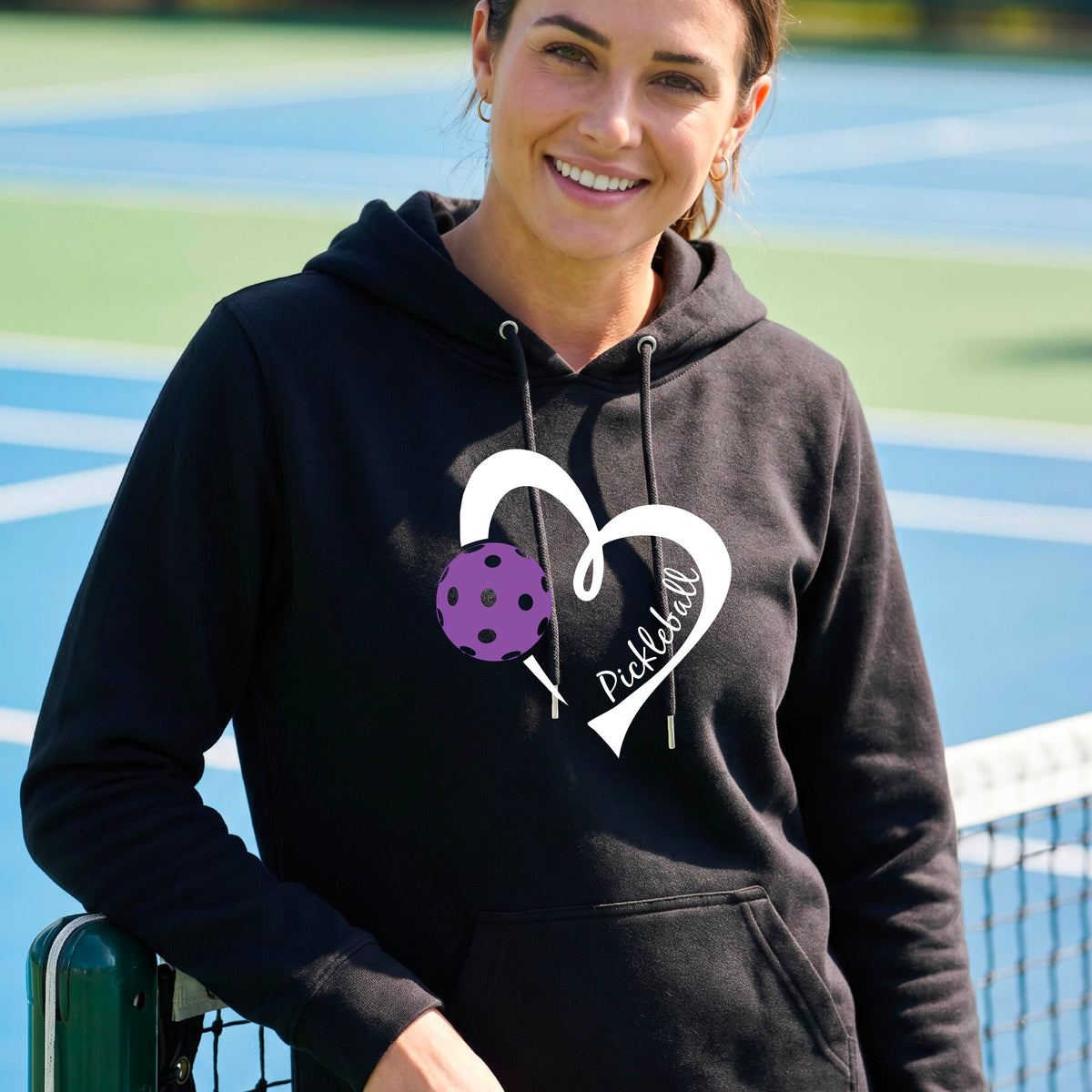 A smiling man wearing a black hoodie with a white heart outline graphic and a purple picklkeball design and the texgt 'Pickleball' on the chest, standing on a pickleball court.
