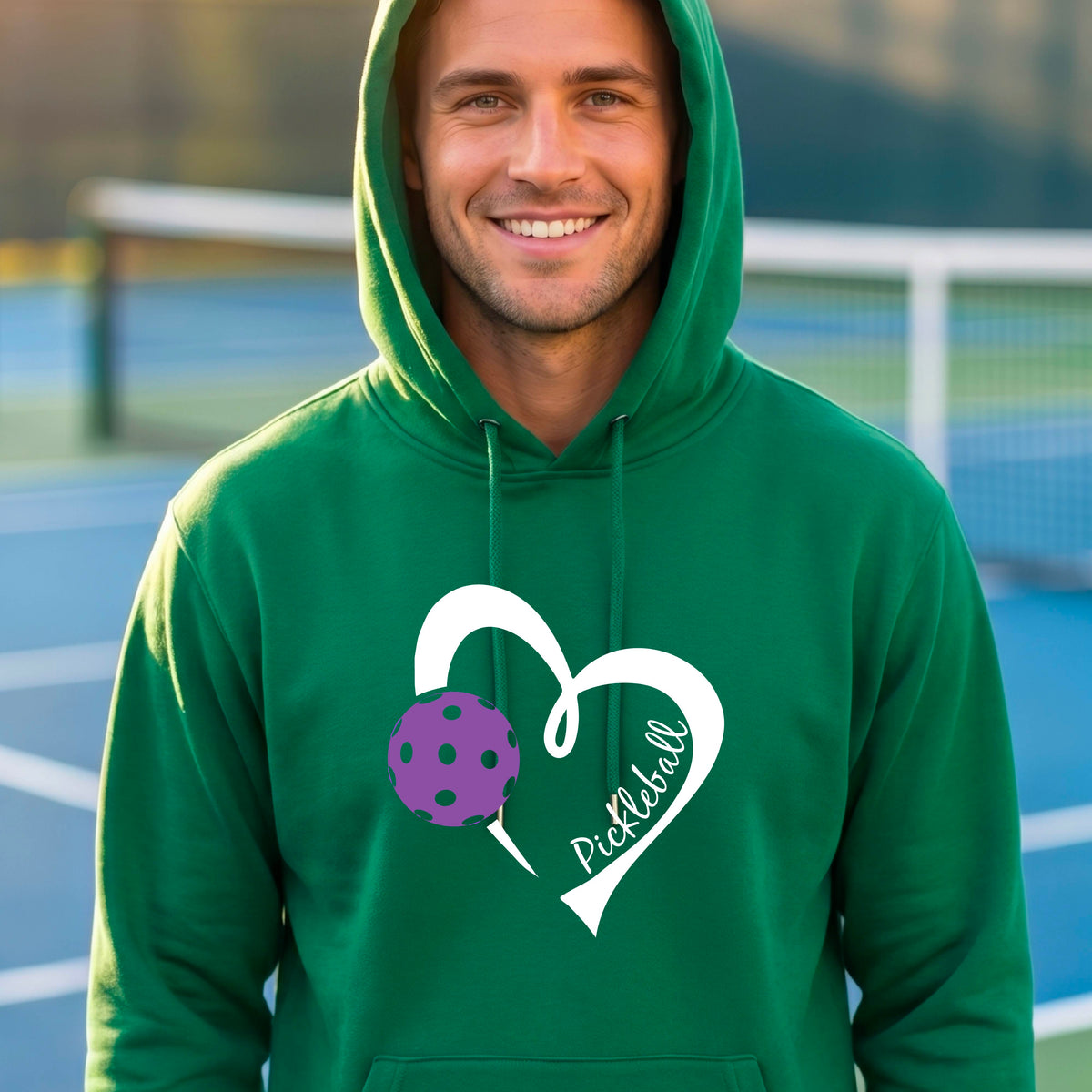 A smiling man wearing a green hoodie with a white heart outline graphic and a purple picklkeball design and the texgt 'Pickleball' on the chest, standing on a pickleball court.