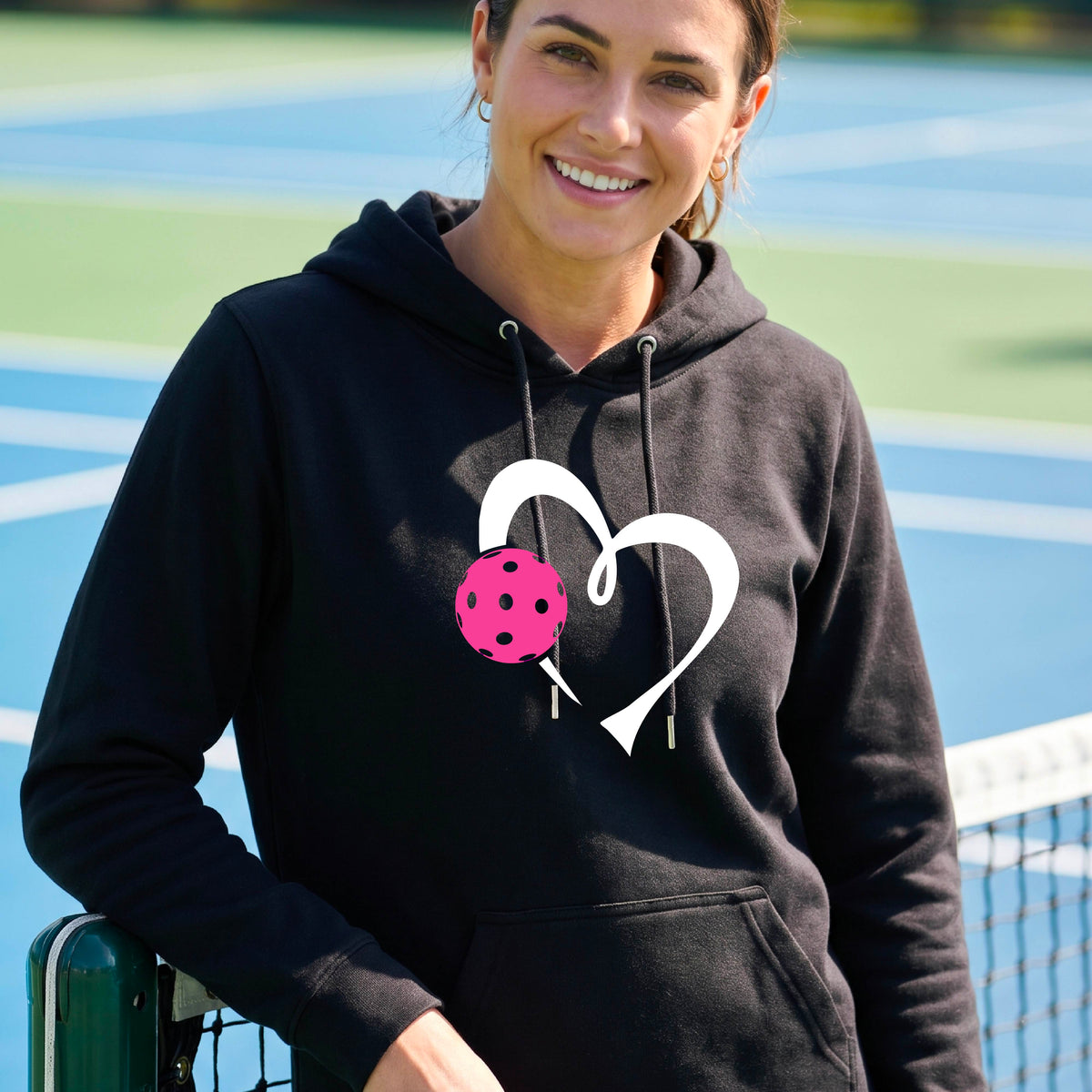 A woman wearing a black unisex hoodie with a white heart and pink pickleball graphic on the front,.
