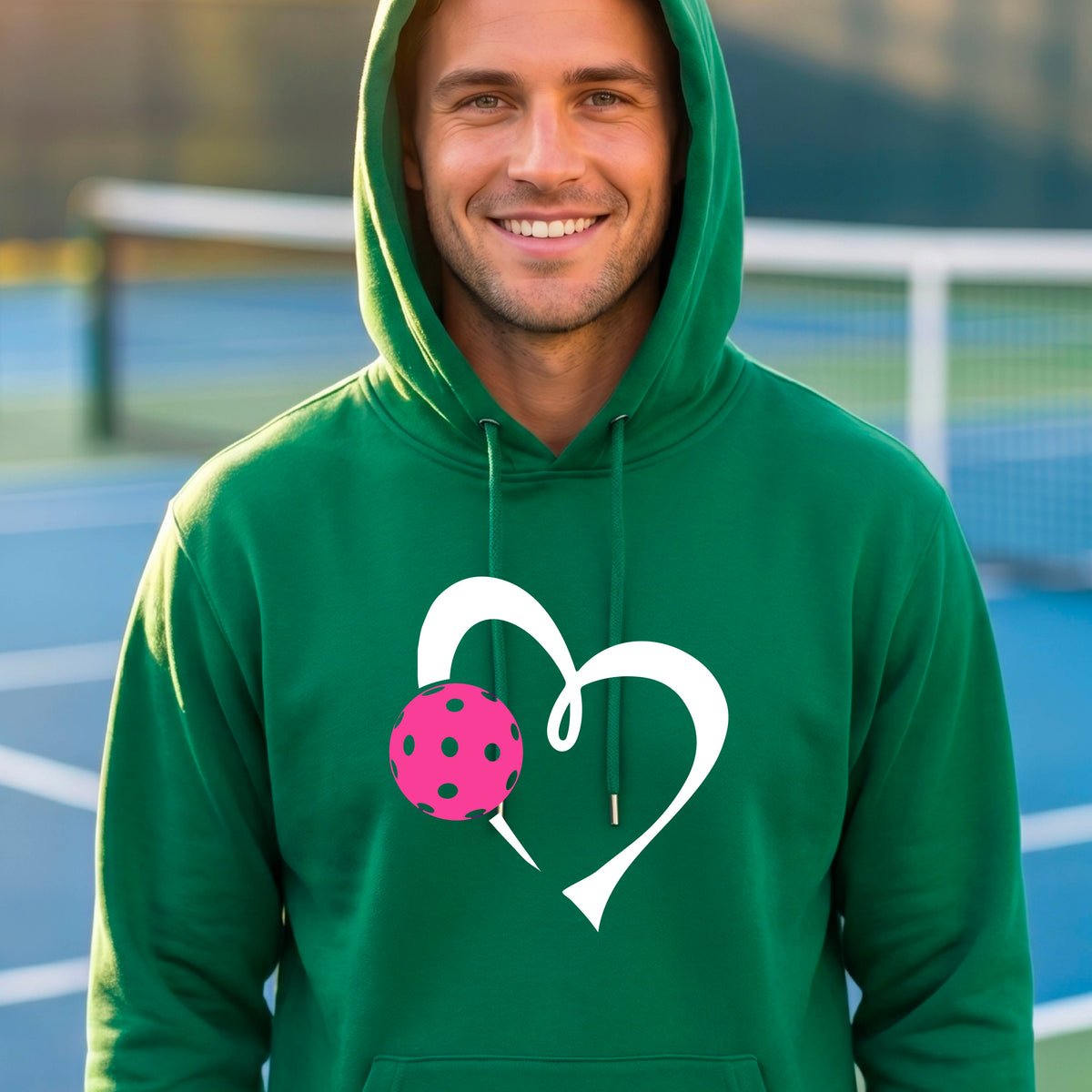 A man wearing a green unisex hoodie with a white heart and pink pickleball graphic on the front,.