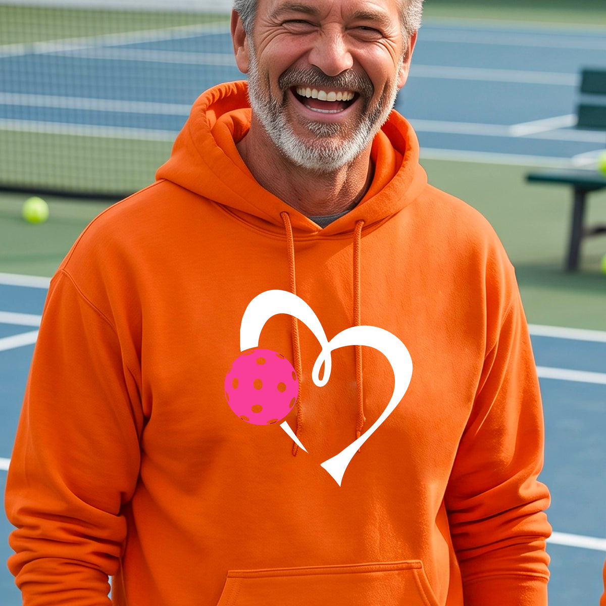 A man wearing an orange unisex hoodie with a white heart and pink pickleball graphic on the front,.
