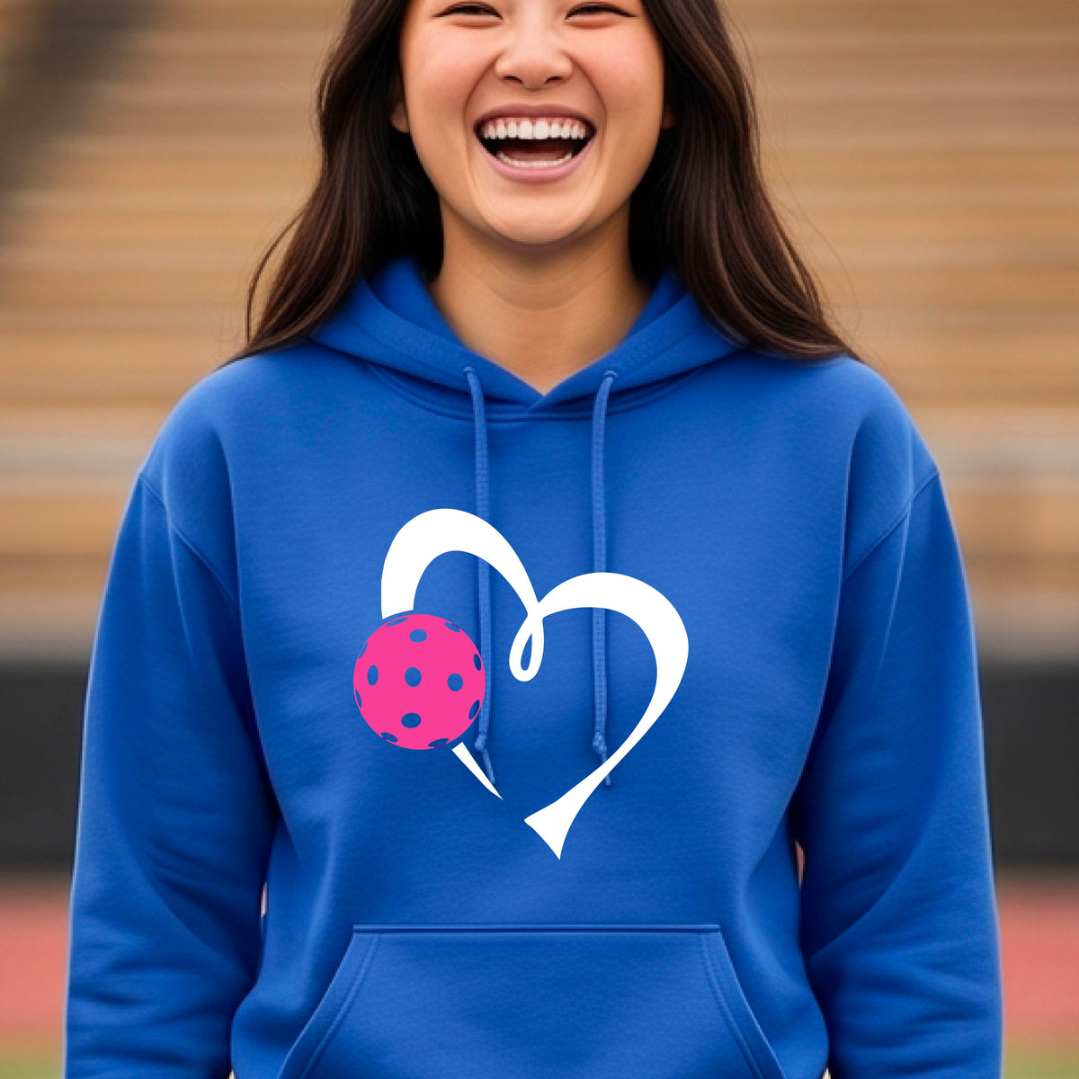 A woman wearing a bright royal blue unisex hoodie with a white heart and pink pickleball graphic on the front,.