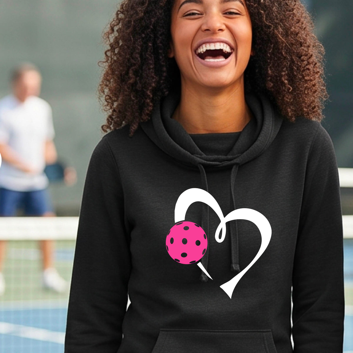 A woman wearing a black cowl-neck hoodie with a white heart outline and a pink pickleball graphic, standing on a pickleball court .