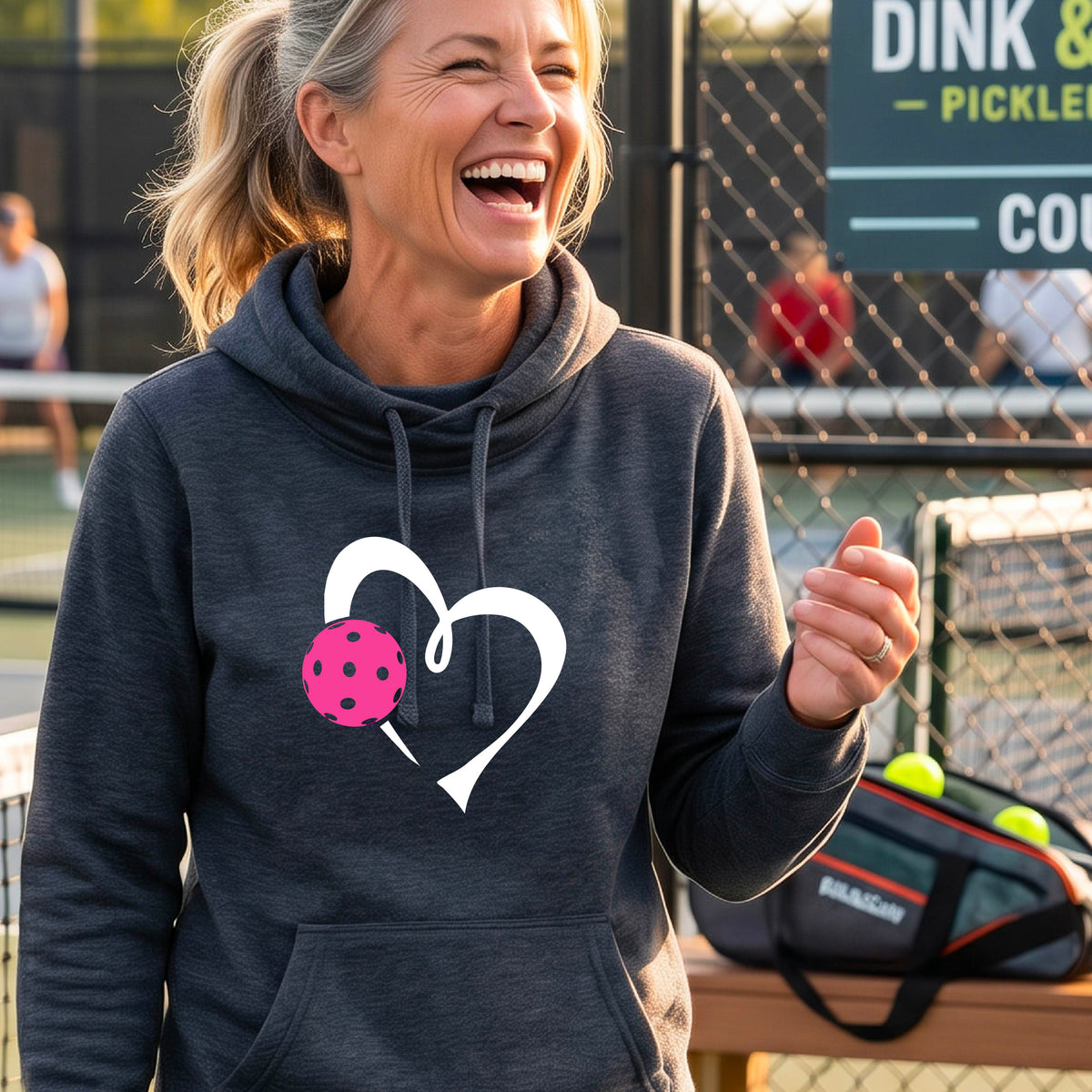A woman wearing a navy blue cowl-neck hoodie with a white heart outline and a pink pickleball graphic, standing on a pickleball court .