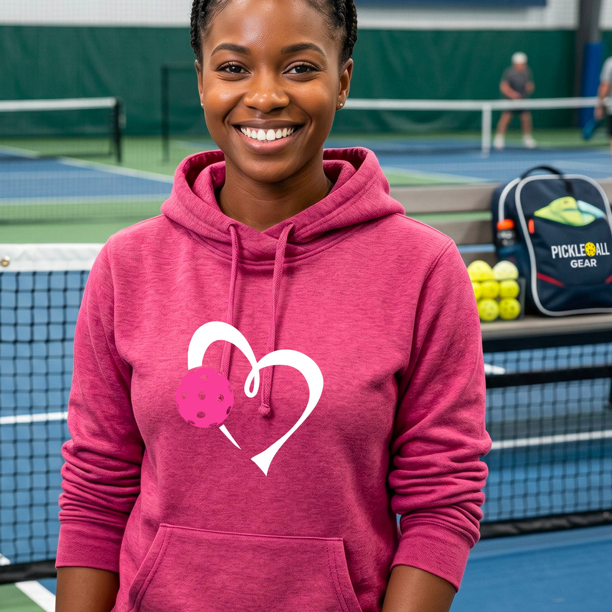 A woman wearing a pink cowl-neck hoodie with a white heart outline and a pink pickleball graphic, standing on a pickleball court .