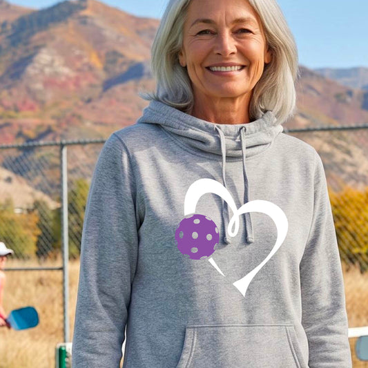 A smiling woman wearing a gray hoodie with a white heart outline and a purple pickleball graphic on the chest. 