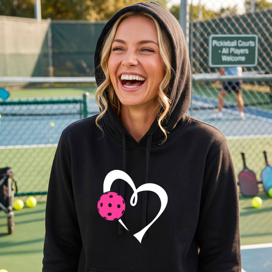 A woman laughing on a pickleball court wearing a black hoodie with a white heart outline and a pink pickleball graphic on the front.