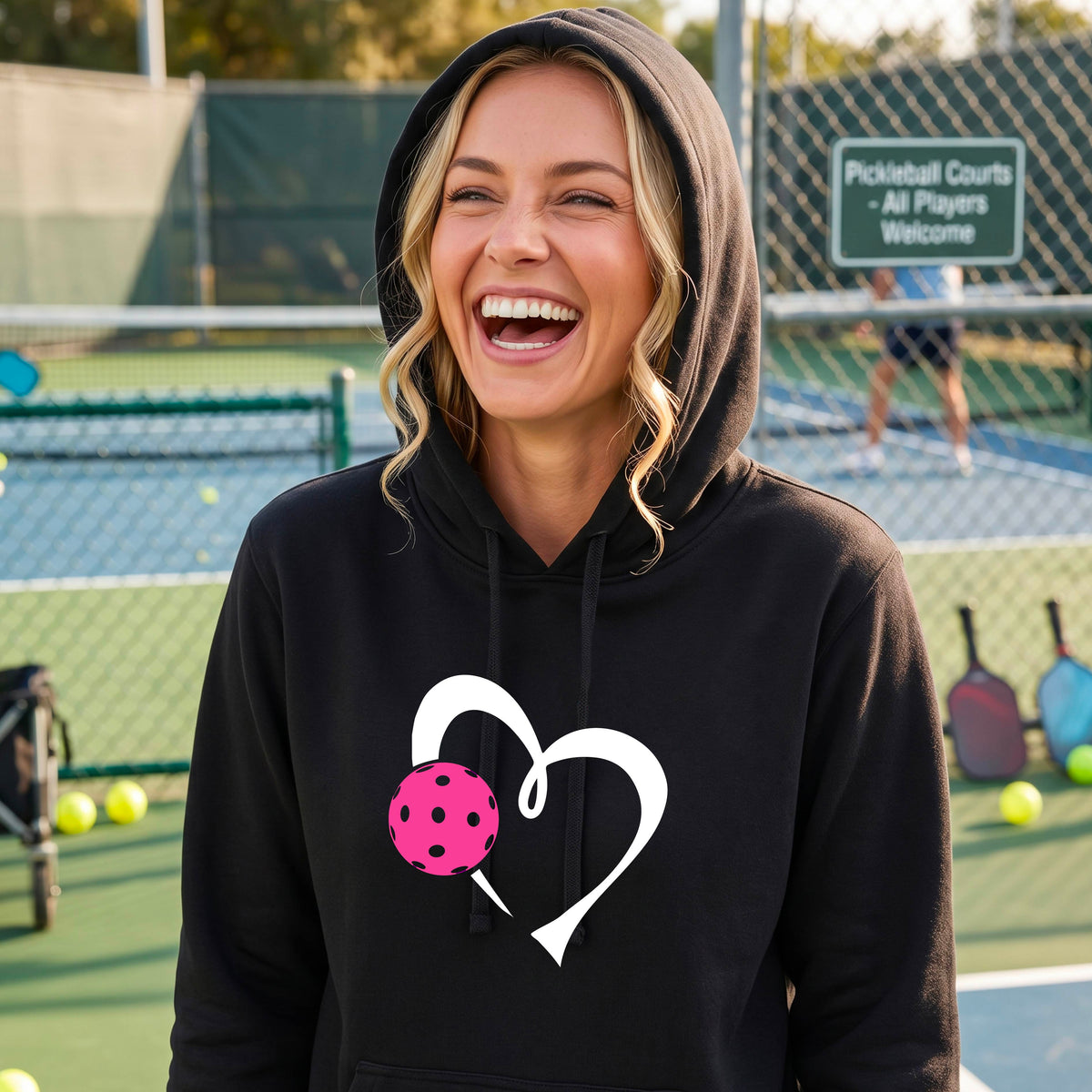 A woman wearing a black hoodie pullover with a white heart and a pink pickleball graphic on the front, standing on a blue pickleball court.