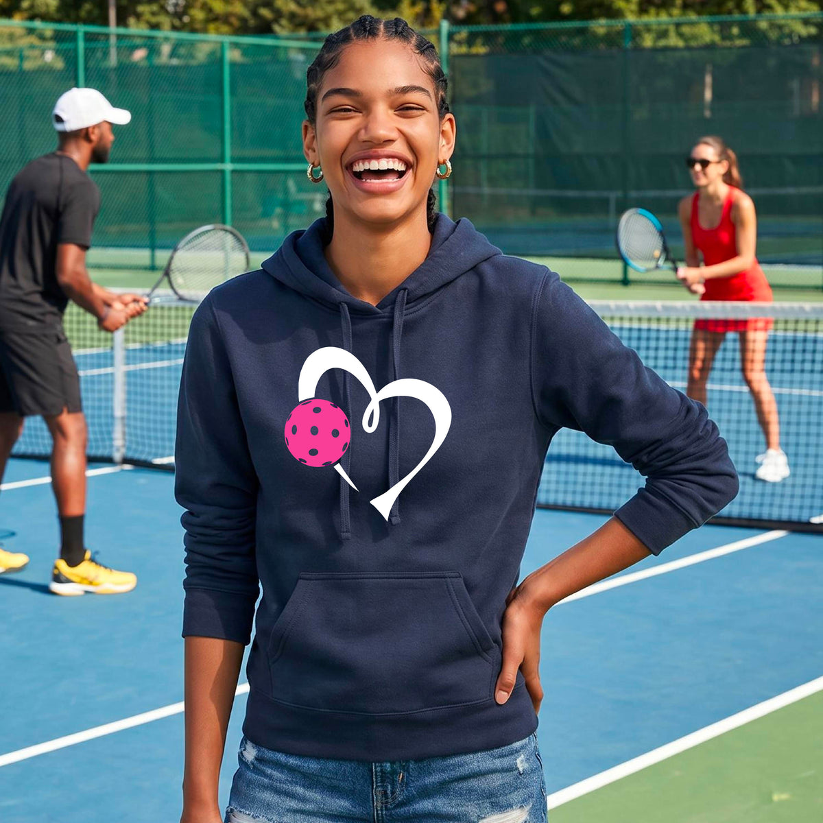 A woman wearing a navy blue hoodie pullover with a white heart and a pink pickleball graphic on the front, standing on a blue pickleball court.