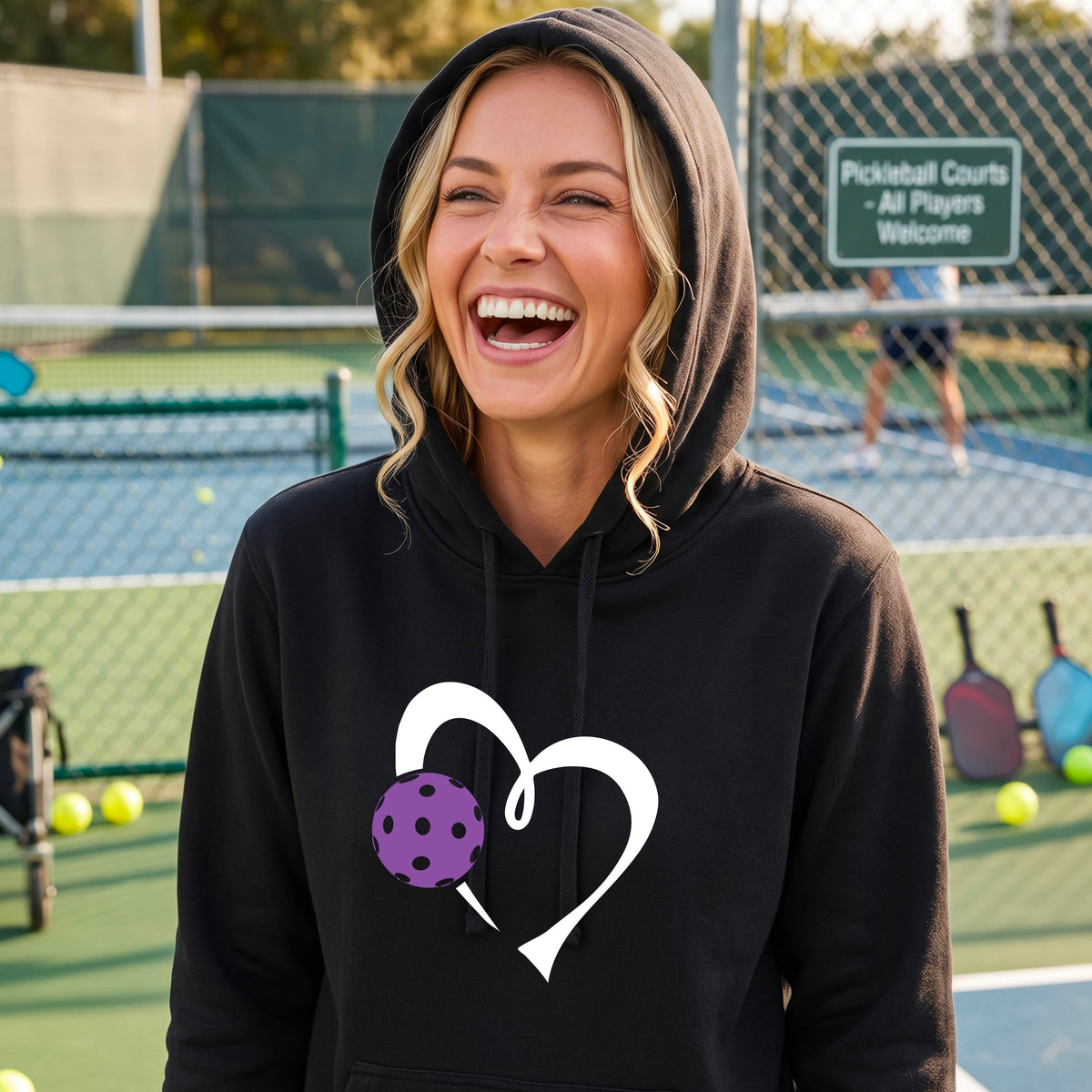 A woman smiling on an outdoor pickleball court wearing a black hoodie with a white heart outline and a purple pickleball graphic on the front. 